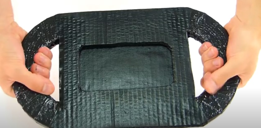 Mientras va secando, empezaremos a hacer la base. Recorta una tira de cartón de unos 8 cm de ancho y dóblala en tres partes, dejando una un par de centímetros más larga que las otras dos. Luego refuérzala añadiendo otras capas de cartón, tal cual como hicimos con el volante. Una vez reforzadas, procedemos a pegar el extremo más corto con el más largo con la cinta americana y algo de pegamento. Una vez todo haya secado correctamente, abrimos un agujero en la parte superior de la más larga.